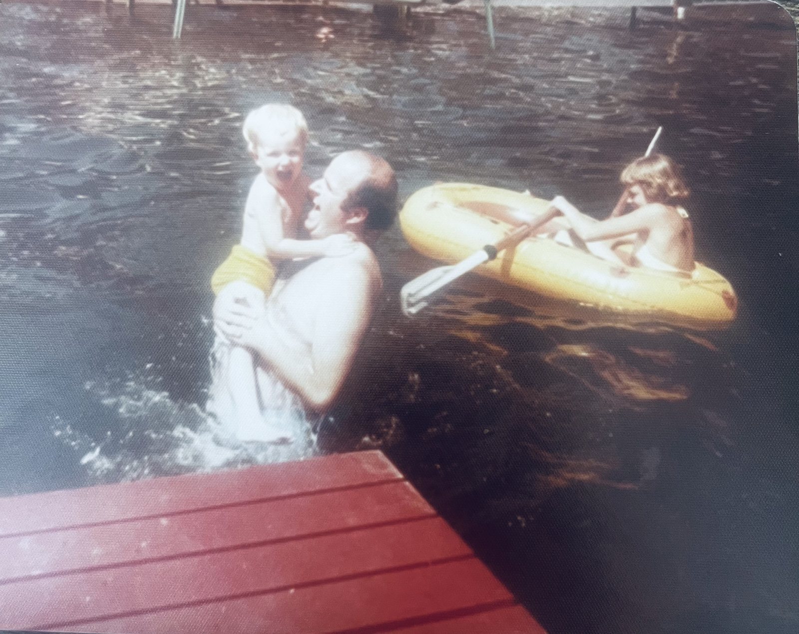 William as a small boy, laughing in his father's arms in a lake beside a red dock, while another child rows a yellow raft nearby